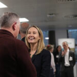 A woman with blonde hair smiling whilst talking to a man in a burgundy jumper at the Ecology annual general meeting 2024.